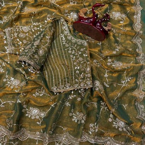 May include: An olive green saree with intricate floral embroidery and a matching blouse. The saree features a scalloped border and is embellished with silver-colored sequins. A decorative maroon figurine sits atop the saree.