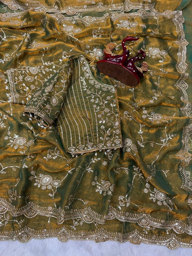 May include: An olive green saree with intricate floral embroidery and a matching blouse. The saree features a scalloped border and is embellished with silver-colored sequins. A decorative maroon figurine sits atop the saree.
