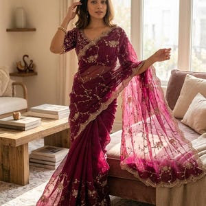 May include: A burgundy saree with intricate gold embroidery and a scalloped border. The sheer fabric drapes elegantly, showcasing floral and vine patterns. The saree features a matching blouse with similar embellishments, creating a cohesive and ornate ensemble.