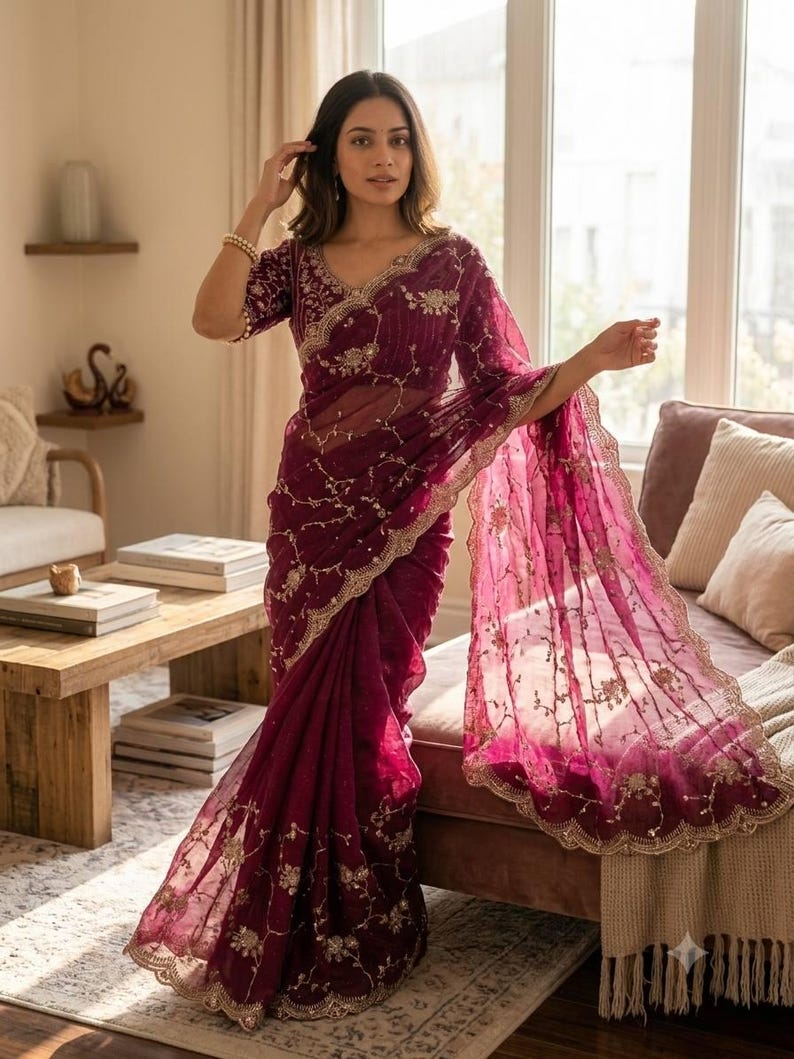 May include: A burgundy saree with intricate gold embroidery and a scalloped border. The sheer fabric drapes elegantly, showcasing floral and vine patterns. The saree features a matching blouse with similar embellishments, creating a cohesive and ornate ensemble.