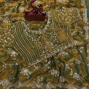 May include: Olive green saree with intricate floral embroidery and sequin embellishments. The saree features a matching blouse with similar detailing. A decorative burgundy and gold figurine sits atop the saree.