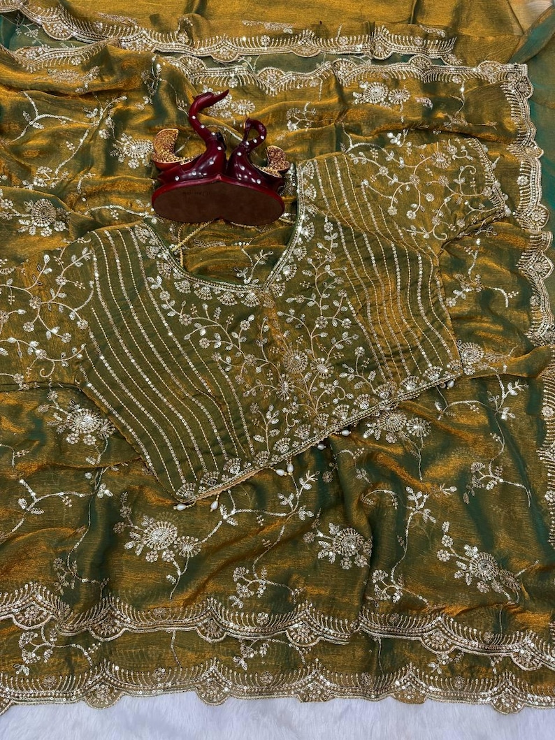 May include: Olive green saree with intricate floral embroidery and sequin embellishments. The saree features a matching blouse with similar detailing. A decorative burgundy and gold figurine sits atop the saree.