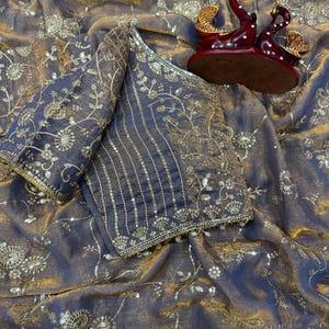 May include: A dark purple and gold sari with intricate floral embroidery and embellishments. The sari is draped over a matching blouse with detailed sleeves. A decorative red and gold swan figurine sits on the fabric.