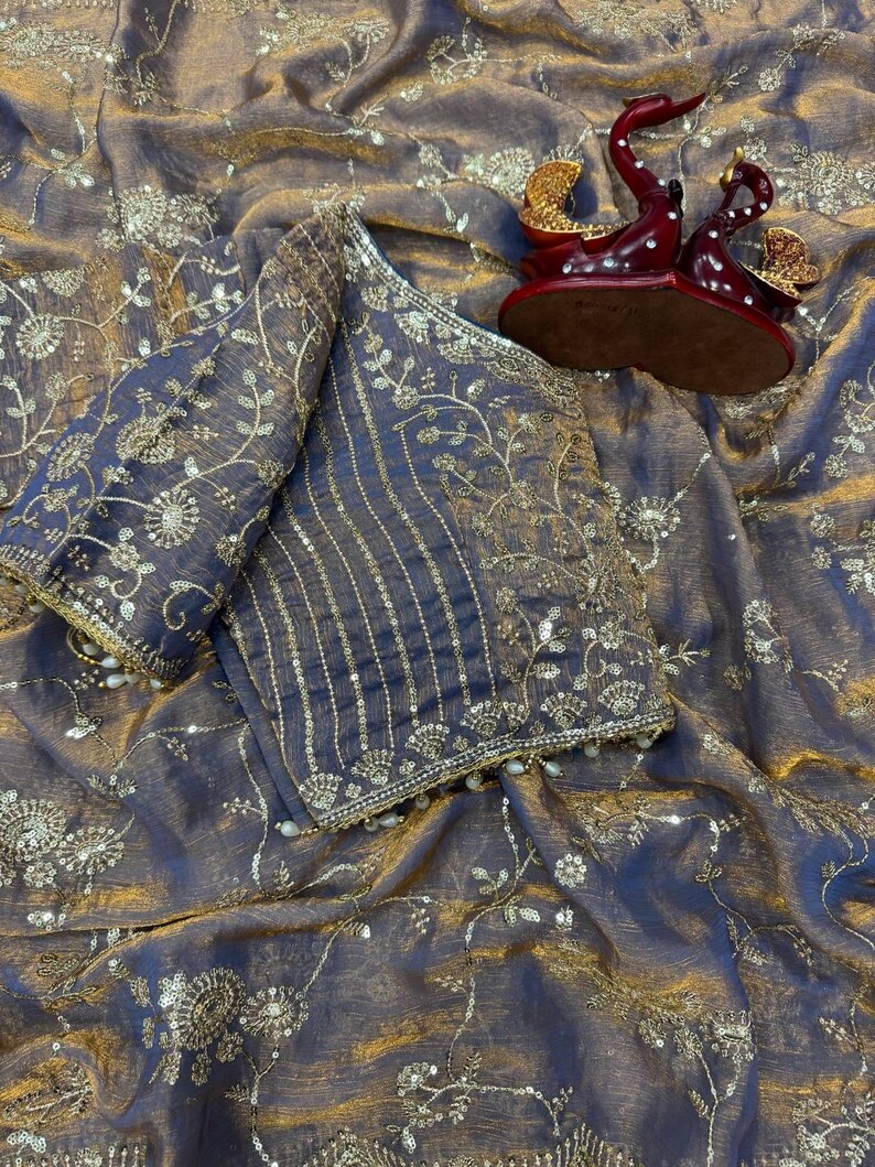 May include: A dark purple and gold sari with intricate floral embroidery and embellishments. The sari is draped over a matching blouse with detailed sleeves. A decorative red and gold swan figurine sits on the fabric.