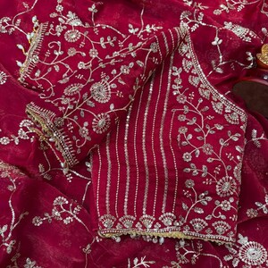 May include: A deep red, embroidered blouse and fabric. The blouse features intricate silver floral embroidery and vertical silver stripes. The fabric has a delicate, flowing texture, with silver floral patterns throughout. The edges are trimmed with gold and small white beads.