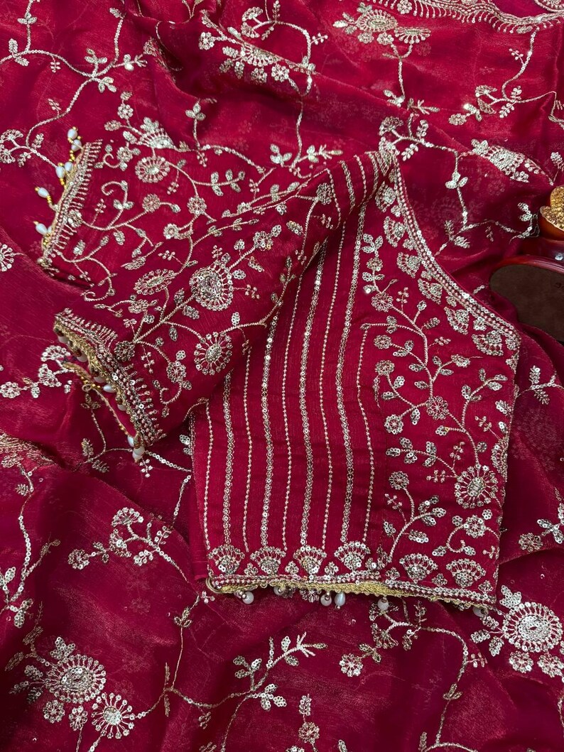May include: A deep red, embroidered blouse and fabric. The blouse features intricate silver floral embroidery and vertical silver stripes. The fabric has a delicate, flowing texture, with silver floral patterns throughout. The edges are trimmed with gold and small white beads.