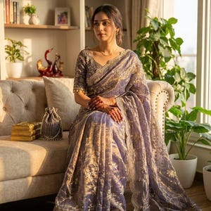 May include: A woman wearing a lavender sari with gold embroidery, seated on a beige sofa. The sari features intricate floral patterns and a scalloped border. The woman is wearing a matching blouse with short sleeves and gold trim.