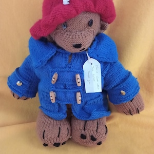 May include: Hand-knitted teddy bear wearing a red hat secured with a safety pin and a blue coat with wooden buttons. The bear has brown fur, black eyes, and a small tag. A charming, handmade toy.