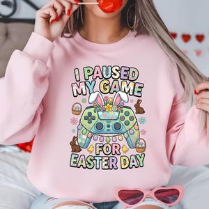 Könnte beinhalten: Ein hellrosa Sweatshirt mit dem Text "I PAUSED MY GAME FOR EASTER DAY". Die Grafik zeigt einen Videospiel-Controller, der mit Osterelementen wie Hasenohren, Blumen, Eiern und Hasen verziert ist.
