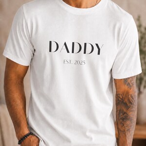 Peut inclure: T-shirt blanc avec le mot "DADDY" en noir et "EST. 2025" en dessous. Le t-shirt est de style col rond. La personne porte un bracelet noir et un jean bleu.