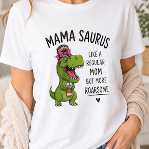 Peut inclure: Un t-shirt blanc avec un motif de dinosaure vert portant un chignon brun et un nœud rose, tenant une tasse de café. Le texte indique MAMA SAURUS LIKE A REGULAR MOM BUT MORE ROARSOME avec une petite icône de cœur noir sous le texte.
