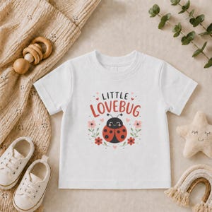 May include: A white short-sleeve t-shirt featuring a graphic of a red and black ladybug surrounded by small pink and red flowers and hearts. The text "LITTLE LOVEBUG" is printed in a dark font above the ladybug illustration on the front.