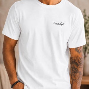 Könnte beinhalten: Weißes T-Shirt mit Rundhalsausschnitt und dem Wort "daddy" in Schreibschrift und einem Herzsymbol. Das Shirt ist einfarbig weiß und hat kurze Ärmel.