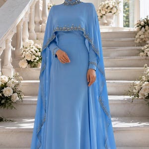 May include: A floor-length light blue gown featuring a high neckline and a matching cape overlay. The collar and the hem of the cape are embellished with intricate silver beadwork and sequins. The dress has long sleeves and a flowing skirt.