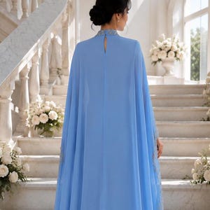 May include: A floor-length light blue gown featuring a high neckline with intricate silver embellishments, a keyhole cutout at the back, and a flowing cape-style overlay that extends from the shoulders to the hem, creating a draped silhouette.