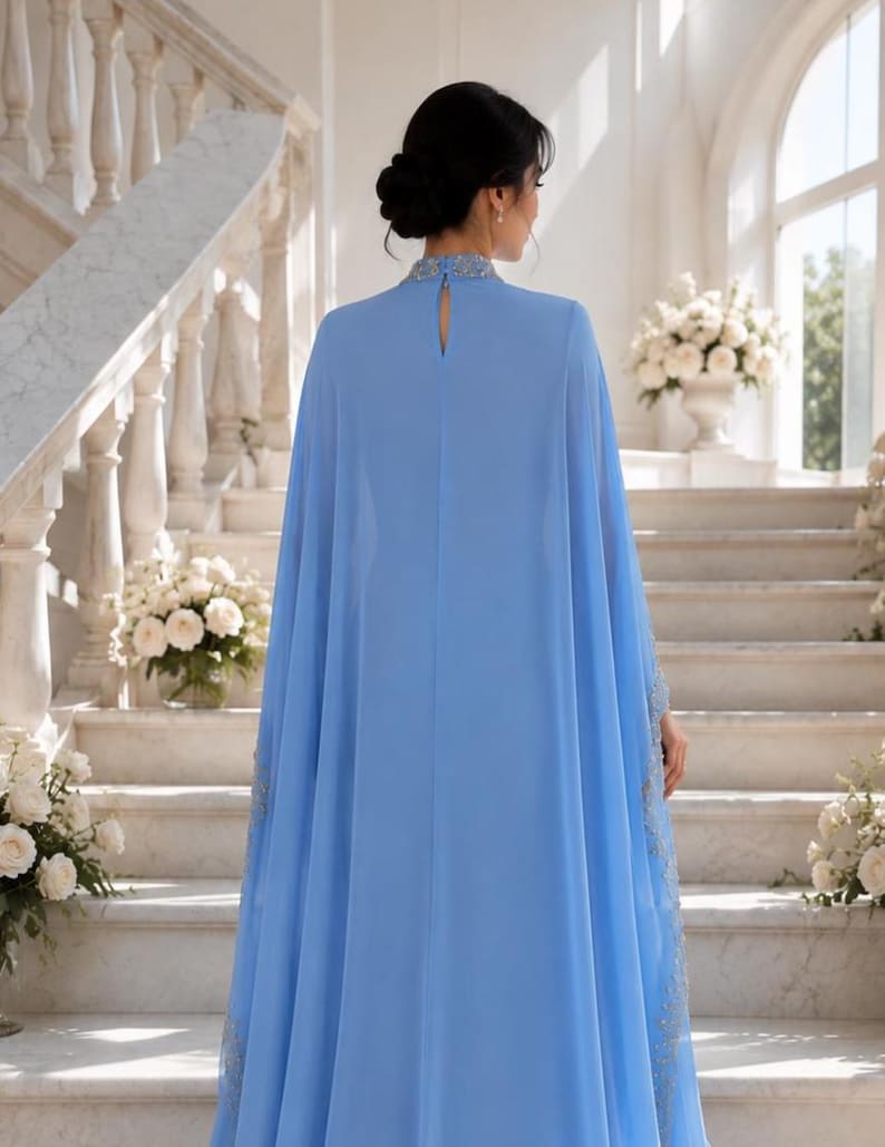 May include: A floor-length light blue gown featuring a high neckline with intricate silver embellishments, a keyhole cutout at the back, and a flowing cape-style overlay that extends from the shoulders to the hem, creating a draped silhouette.
