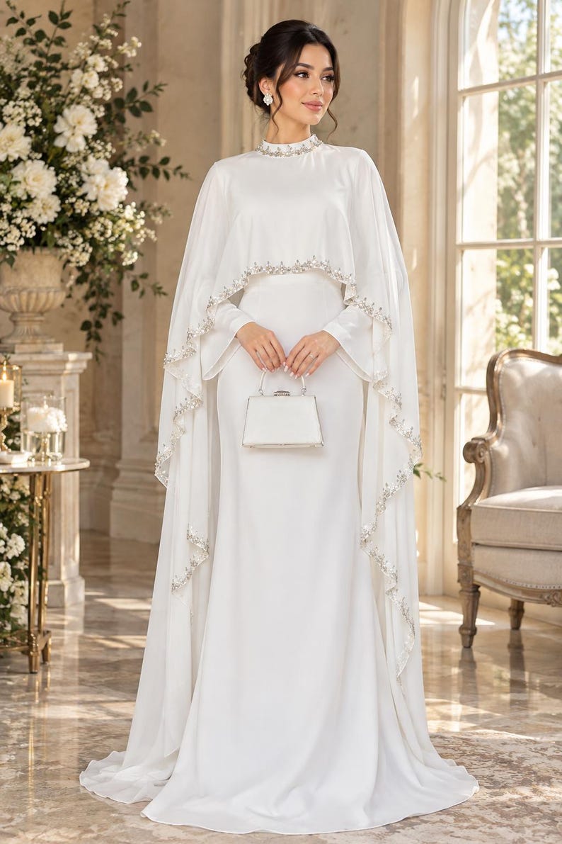 May include: A floor-length white gown featuring a high neckline and a flowing cape overlay. The collar and the scalloped edges of the cape are embellished with intricate silver beadwork and crystals. A small, structured white handbag is held in front.
