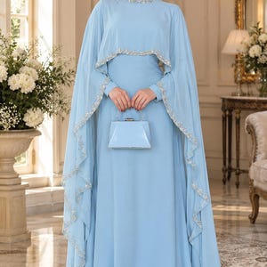 May include: A floor-length light blue gown featuring a high neckline and a matching cape overlay. The neckline and the edges of the cape are embellished with silver beads and clear crystals. A small, structured light blue clutch bag is held.