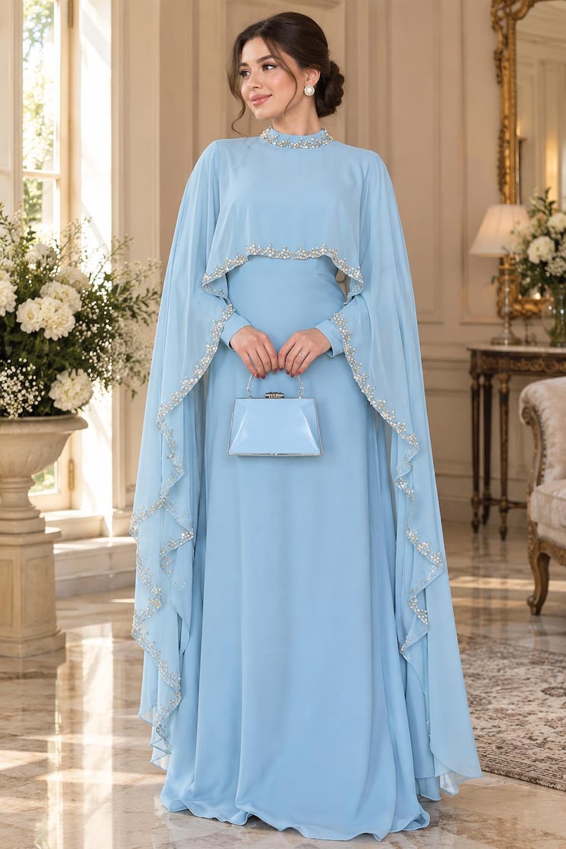 May include: A floor-length light blue gown featuring a high neckline and a matching cape overlay. The neckline and the edges of the cape are embellished with silver beads and clear crystals. A small, structured light blue clutch bag is held.