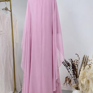 May include: A floor-length, light pink, sleeveless gown featuring a high neckline adorned with silver-toned embellishments and a keyhole opening at the back. The dress is crafted from a flowing, sheer fabric that drapes into wide, cape-like sleeves.