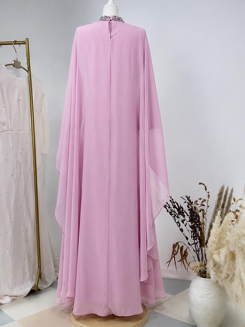 May include: A floor-length, light pink, sleeveless gown featuring a high neckline adorned with silver-toned embellishments and a keyhole opening at the back. The dress is crafted from a flowing, sheer fabric that drapes into wide, cape-like sleeves.