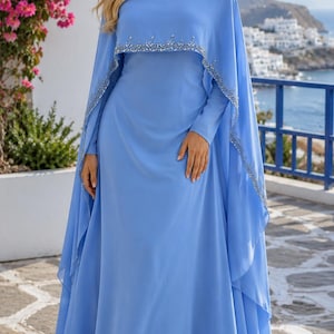 May include: A floor-length light blue gown featuring a high neckline and a cape-style overlay. The collar and the scalloped edge of the cape are embellished with silver beads and crystals. The dress has long sleeves and a flowing, full-length skirt.