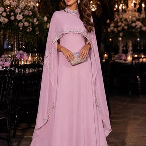 May include: A floor-length lavender gown featuring a high neckline and an attached cape overlay. The collar and the curved hem of the cape are embellished with silver beadwork and sequins. The dress is paired with a small, rectangular silver clutch.