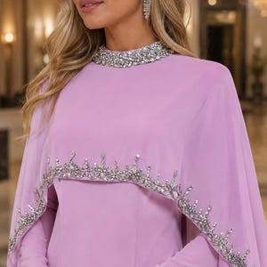 May include: A light purple long-sleeved dress featuring a high neckline adorned with clear crystals and a matching cape overlay. The cape is embellished with a decorative border of silver-toned crystals and beads along the scalloped lower edge.