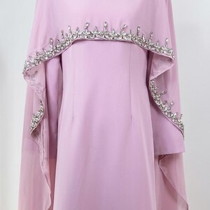 May include: A light lavender floor-length dress featuring a high neckline adorned with clear crystals. The garment includes a matching sheer cape overlay that drapes over the shoulders, embellished with a border of sparkling clear crystals.
