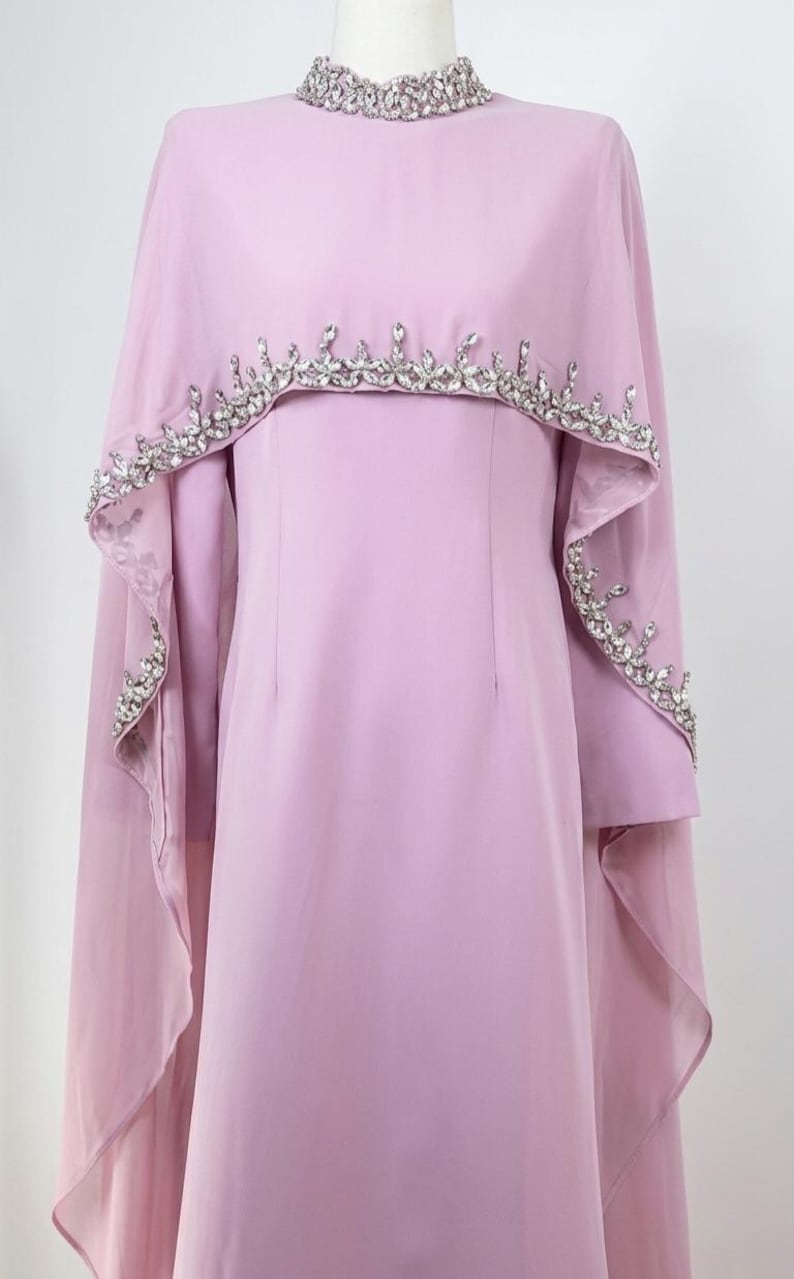 May include: A light lavender floor-length dress featuring a high neckline adorned with clear crystals. The garment includes a matching sheer cape overlay that drapes over the shoulders, embellished with a border of sparkling clear crystals.