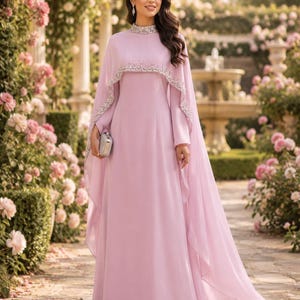 May include: A floor-length light purple gown featuring a high neckline and a cape-style overlay. The collar and the edge of the cape are embellished with silver beads and crystals. The dress has long sleeves and is paired with a small silver clutch.