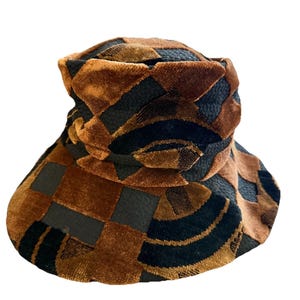 May include: A bucket hat featuring a geometric pattern in shades of dark brown velvet and black textured fabric. The hat has a rounded crown and a wide, downward-sloping brim, with the velvet and textured panels alternating across the entire surface.