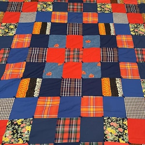 Puede incluir: Un edredón de retazos con una vibrante mezcla de colores y patrones. El edredón presenta cuadrados de telas rojas, azules, a cuadros, florales y de mezclilla. El diseño es una cuadrícula de varios cuadrados de tela, creando un textil acogedor y colorido.