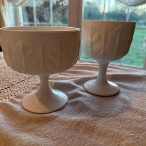 Peut inclure: Deux verres à dessert blancs à pied, ornés d'un motif texturé en forme de feuilles. Les verres ont un large bol et un pied en forme de socle, posés sur une nappe en lin de couleur neutre. L'arrière-plan comprend une fenêtre.