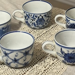 May include: Five white ceramic teacups with blue floral and geometric patterns. Each cup has a white handle and a blue rim. The cups are arranged on a cream-colored, lace-like surface.