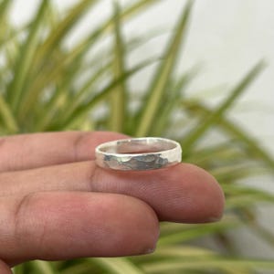 Könnte beinhalten: Ein silberner Ring mit gehammerter Textur. Der Ring ist rund und hat eine leicht unregelmäßige Oberfläche. Der Ring wird in der Hand einer Person gehalten, mit einem verschwommenen grün-gelben Hintergrund.