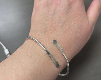 Puede incluir: Un brazalete de color plateado. El diseño abierto presenta dos extremos planos, ligeramente curvados. El brazalete se lleva en la muñeca, con un fondo gris visible.