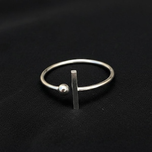 Könnte beinhalten: Ein minimalistischer Silberring mit einem kleinen Ball und einem vertikalen Strich-Design. Der Ring ist vor einem schwarzen Hintergrund platziert, was seine schlichte, moderne Ästhetik unterstreicht. Der Ring hat ein dünnes, kreisförmiges Band.