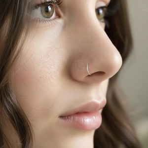 Puede incluir: Primer plano de un rostro, destacando un pequeño aro de nariz plateado. El aro es un círculo fino y sencillo. La persona tiene piel clara, cabello castaño y ojos verdes. La imagen está bien iluminada y enfocada.