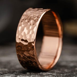 Könnte beinhalten: Ein roségoldener Ring mit gehämmertem Muster. Der Ring hat ein breites Band und eine polierte Innenseite. Die gehämmerten Textur erzeugt eine einzigartige Oberfläche. Der Ring wird auf einer dunklen, strukturierten Oberfläche präsentiert.