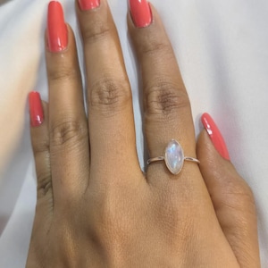 May include: A silver ring featuring a marquise-cut, iridescent gemstone. The ring is set on a thin band. The hand wearing the ring has coral-coloured nail polish.