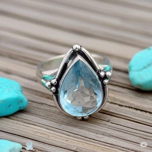 Könnte beinhalten: Ein silberner Ring mit einem großen, tropfenförmigen, hellblauen Edelstein. Der Ring hat eine detaillierte Fassung mit kleinen, runden Akzenten und liegt auf einer Holzoberfläche. Der Edelstein ist facettiert.
