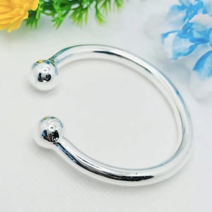 Könnte beinhalten: Ein silberfarbenes, offenes Armband mit glattem, abgerundetem Design. Das Armband hat an jedem Ende einen kugelförmigen Akzent. Der Schmuck wird auf einer weißen Oberfläche mit floralen Akzenten im Hintergrund präsentiert.