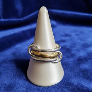 Könnte beinhalten: Ein silber- und goldfarbener Ring mit drei ineinandergreifenden Bändern. Der Ring wird auf einem weißen, kegelförmigen Ständer vor blauem Hintergrund präsentiert. Das Design des Rings zeigt ein goldenes Band zwischen zwei silbernen Bändern.