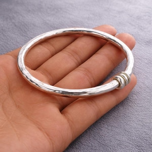 Könnte beinhalten: Ein silbernes Armreif-Armband mit gehämmertem Muster. Das Armband hat ein schlichtes, elegantes Design mit runder Form. Der Verschluss hat drei kleine, goldfarbene Ringe. Dieses Schmuckstück ist ein stilvolles Accessoire.