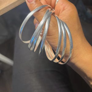 Könnte beinhalten: Eine Hand hält mehrere silberfarbene Armreifen. Die Armreifen haben eine glatte, gebogene Form und eine Oberfläche aus gebürstetem Metall. Das Licht reflektiert sich auf der Oberfläche und erzeugt Glanzlichter. Schlichtes, elegantes Schmuckdesign.