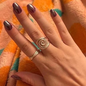 Könnte beinhalten: Eine Hand mit zwei silbernen Ringen, einer mit einem Spiraldesign und der andere mit einem verdrehten Band. Die Nägel sind mit einem metallisch-violetten Lack lackiert. Der Hintergrund ist ein verschwommener orange-pfirsichfarbener Stoff.