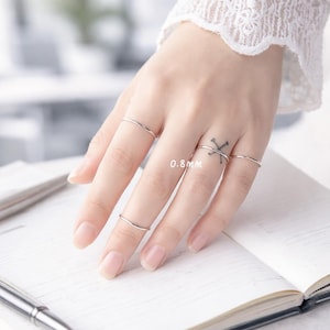 Könnte beinhalten: Eine Hand, die mehrere dünne, silberne Ringe zeigt. Die Ringe sind schlichte Bänder mit einem Durchmesser von 0,8 mm. Die Hand liegt auf einem offenen Notizbuch, ein Stift ist zu sehen. Der Hintergrund ist unscharf.