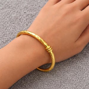 Könnte beinhalten: Ein goldfarbenes Armreif-Armband mit gehämmertem Muster. Das Armband hat ein schlichtes Design mit einem knotenartigen Detail in der Mitte. Das Schmuckstück wird am Handgelenk einer Person gezeigt.