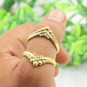 May include: Two gold-coloured rings are shown on a finger. The rings have a V-shape design, with small, round beads along the edges and at the top of the V. The background is blurred, with green and white tones.